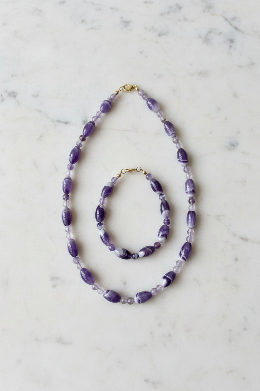 Aurelle Amethyst Schmuckset — Schmuckset aus Amethyst‑Halskette und Armband mit ovalen und runden violetten Naturstein‑Perlen und goldfarbenen Details, auf hellem Stoff minimalistisch arrangiert.