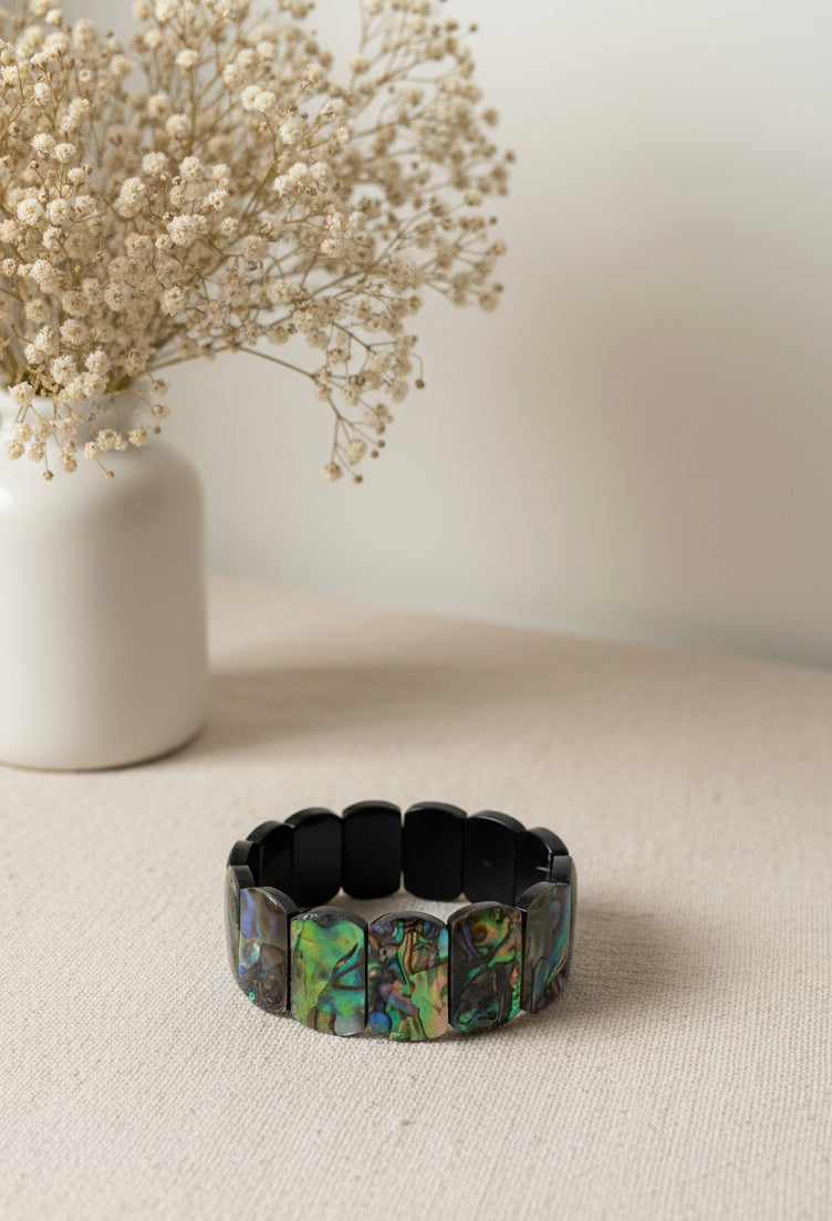 Oceana Meeropal Armband — Irideszierendes Meeropal‑Armband mit rechteckigen Perlen in schimmernden Blau‑ und Grüntönen, minimalistisch auf hellem Stoff neben einer Vase mit Trockenblumen arrangiert.