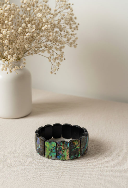Oceana Meeropal Armband — Irideszierendes Meeropal‑Armband mit rechteckigen Perlen in schimmernden Blau‑ und Grüntönen, minimalistisch auf hellem Stoff neben einer Vase mit Trockenblumen arrangiert.