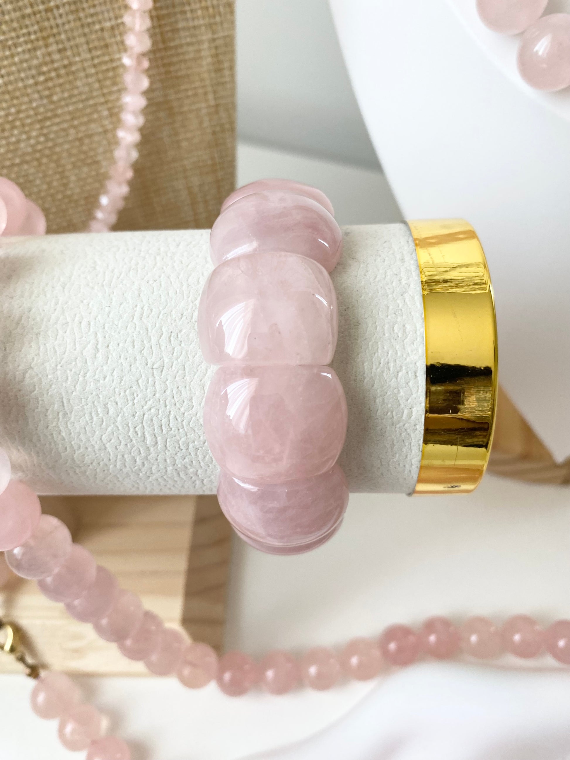 Elastisches Rosenquarz‑Armband aus weich gerundeten roséfarbenen Steinen, präsentiert auf einem weißen texturierten Schmuckdisplay mit goldfarbenem Abschluss. Im Hintergrund liegen passende Rosenquarz‑Ketten auf einem beigen Stoffdisplay und einer weißen Büste.