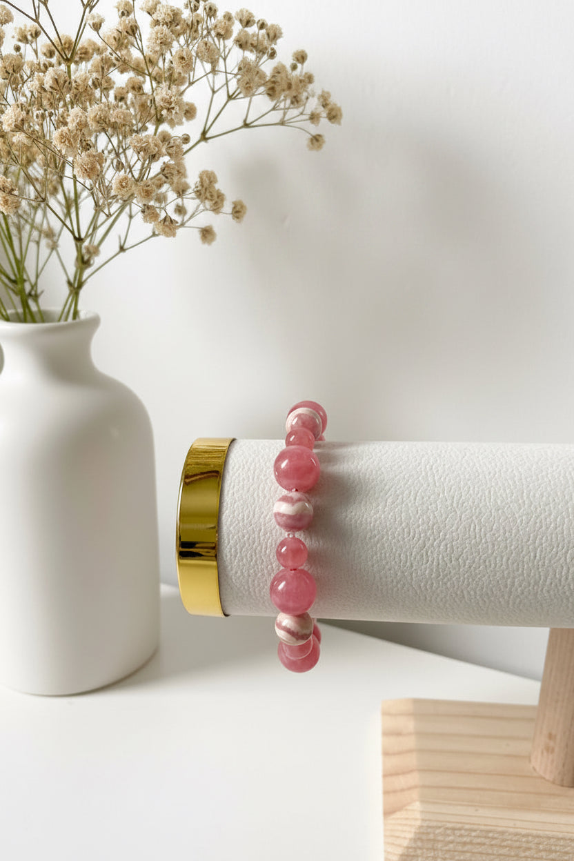 Rhodochrosit‑Armband aus roséfarbenen, leicht gemaserten Edelsteinperlen, präsentiert auf einem weißen zylindrischen Schmuckdisplay mit goldfarbenem Abschluss. Dahinter steht eine weiße Vase mit zarten, beigefarbenen Trockenblumen.