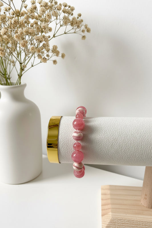 Rhodochrosit‑Armband aus roséfarbenen, leicht gemaserten Edelsteinperlen, präsentiert auf einem weißen zylindrischen Schmuckdisplay mit goldfarbenem Abschluss. Dahinter steht eine weiße Vase mit zarten, beigefarbenen Trockenblumen.
