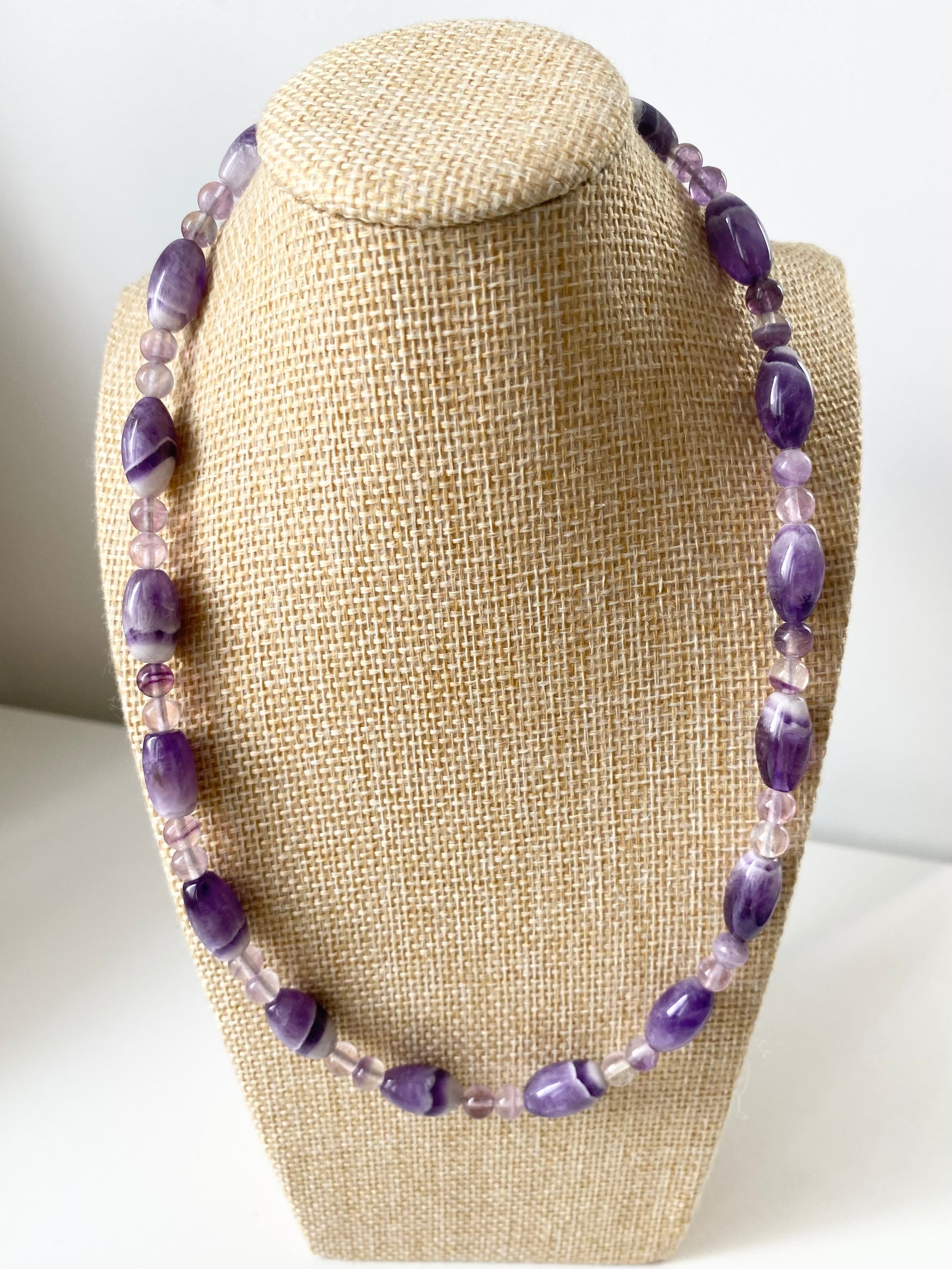 Aurelle Amethyst Halskette — Violette Amethyst‑Halskette mit ovalen und runden Naturstein‑Perlen in verschiedenen Purpurtönen, präsentiert auf einem beigen Schmuckbüsten‑Display.
