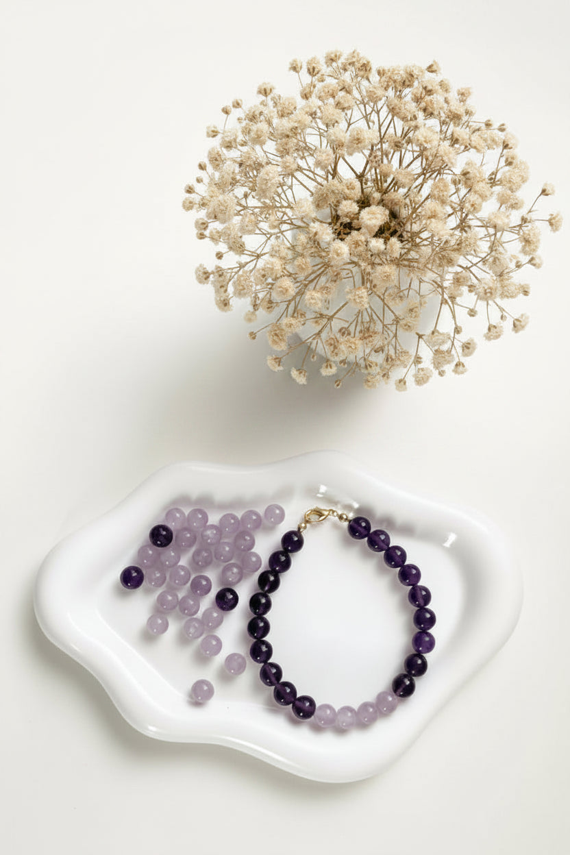 Armband aus runden, violetten Amethystperlen mit goldfarbenem Verschluss, präsentiert auf einer weißen Keramikschale. Daneben liegen einzelne Amethystperlen, darüber ein zarter Zweig getrockneter beige‑farbener Blüten.