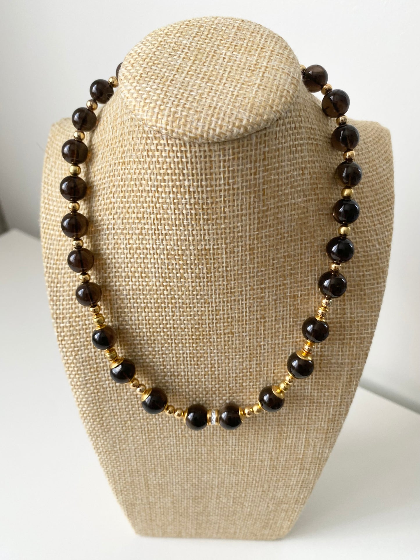 Marione Noir Rauchquarz Halskette — Dunkle Rauchquarz‑Halskette mit runden Naturstein‑Perlen und goldfarbenen Abstandselementen, präsentiert auf einer beigen Schmuckbüste in minimalistischem Stil.