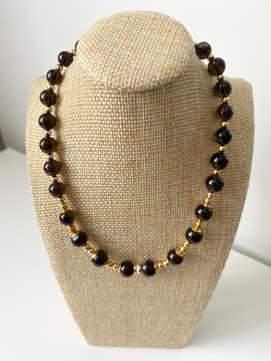 Marione Noir Rauchquarz Halskette — Dunkle Rauchquarz‑Halskette mit runden Naturstein‑Perlen und goldfarbenen Abstandselementen, präsentiert auf einer beigen Schmuckbüste in minimalistischem Stil.