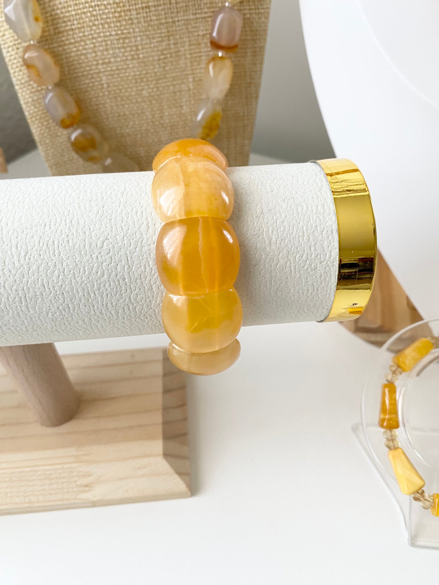 Aragonit‑Armband aus großen, warm gelb‑orangefarbenen Edelsteinperlen, präsentiert auf einem weißen texturierten Schmuckdisplay mit goldfarbenen Endkappen. Dahinter stehen passende Aragonit‑Schmuckstücke auf Stoff‑ und Acrylständern in sanften Naturtönen.