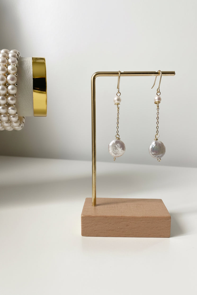 Merelle Pearl Margaux Süsswasserohrringe aus Süsswasserperlen mit kleiner Rundperle und grosser barocker Perle an goldener Fassung, präsentiert auf minimalistischem Schmuckständer.