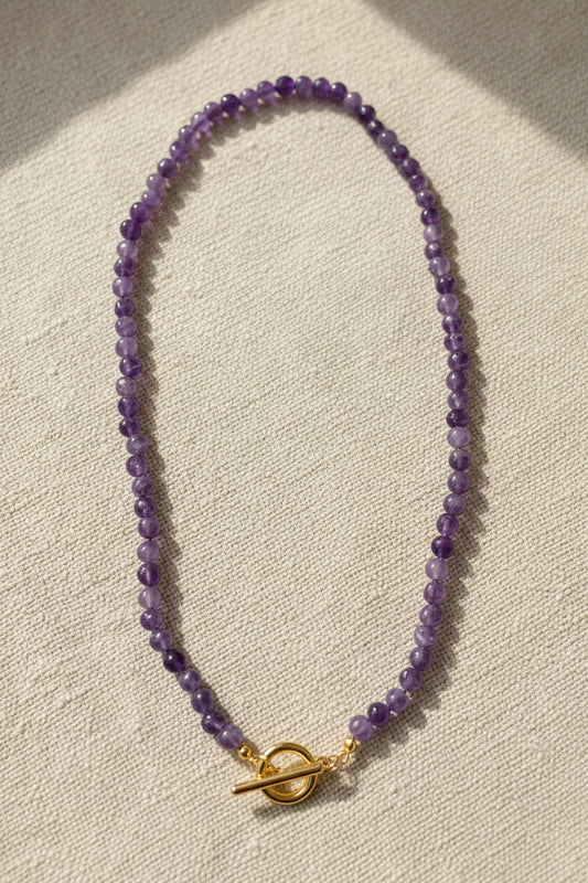Amethyst‑Halskette aus kleinen, polierten violetten Perlen mit goldfarbenem Toggle‑Verschluss, auf beigem Stoff in natürlichem Licht.
