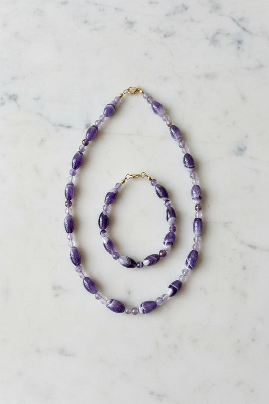 Aurelle Amethyst Schmuckset — Schmuckset aus Amethyst‑Halskette und Armband mit ovalen und runden violetten Naturstein‑Perlen und goldfarbenen Details, auf hellem Stoff minimalistisch arrangiert.