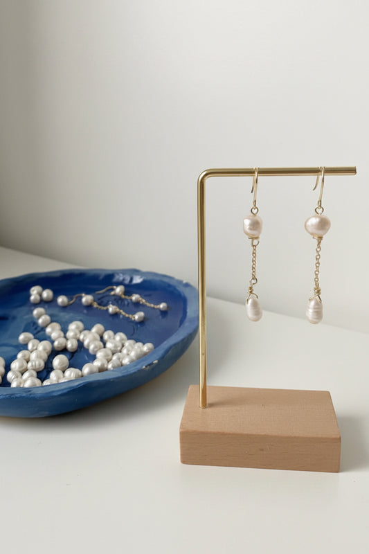 Paar handgefertigter Ohrringe mit zwei barocken Süsswasserperlen, verbunden durch eine 18K gold plated Kette und 14K gold filled Komponenten, präsentiert auf einem minimalistischen Ständer mit Holzsockel; im Hintergrund lose Perlen in einer blauen Schale.