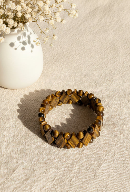 Tigerauge Armband Tara mit braun‑gold schimmernden, rund und quadratisch geschliffenen Perlen auf hellem Stoffhintergrund neben einer kleinen Vase mit Trockenblumen.