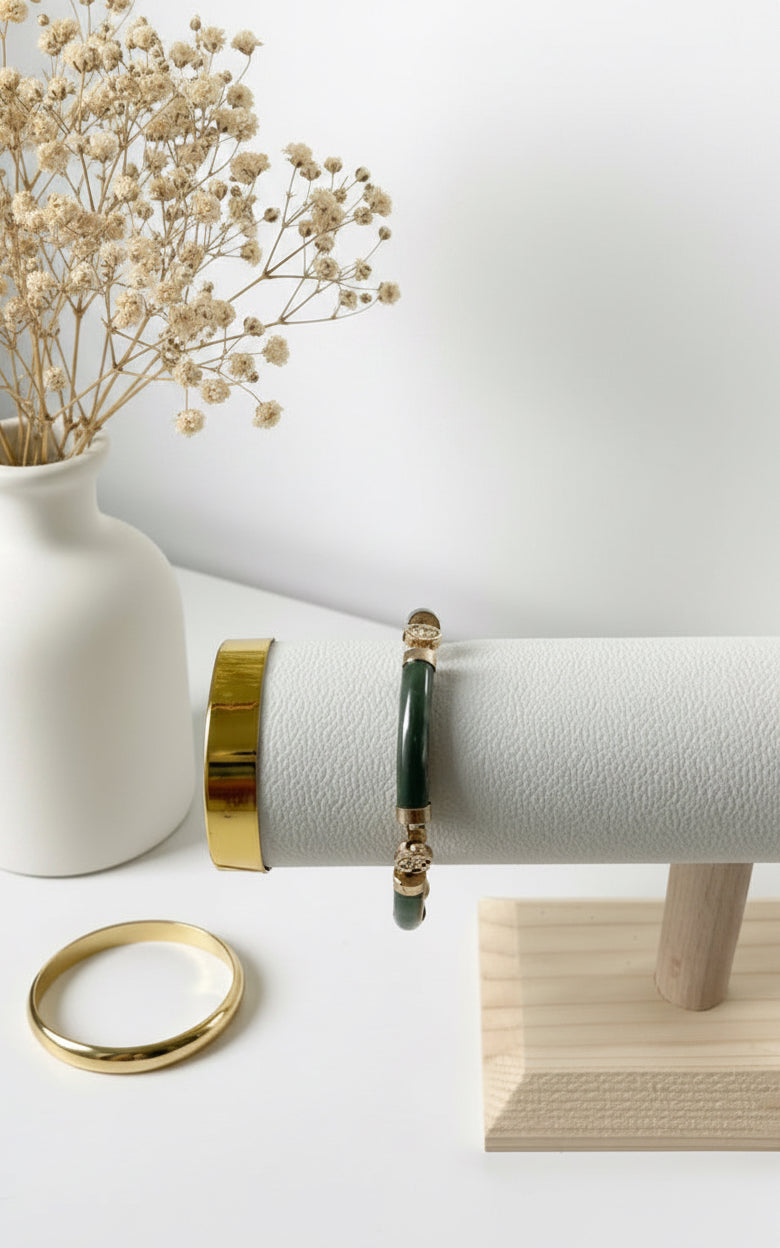 Jade‑Armband mit tiefgrünen Edelsteinperlen und goldfarbenen Akzenten, präsentiert auf einem weißen, texturierten Schmuckständer aus hellem Holz. Daneben liegt ein goldfarbener Armreif; im Hintergrund steht eine weiße Vase mit getrockneten beigefarbenen Blüten.